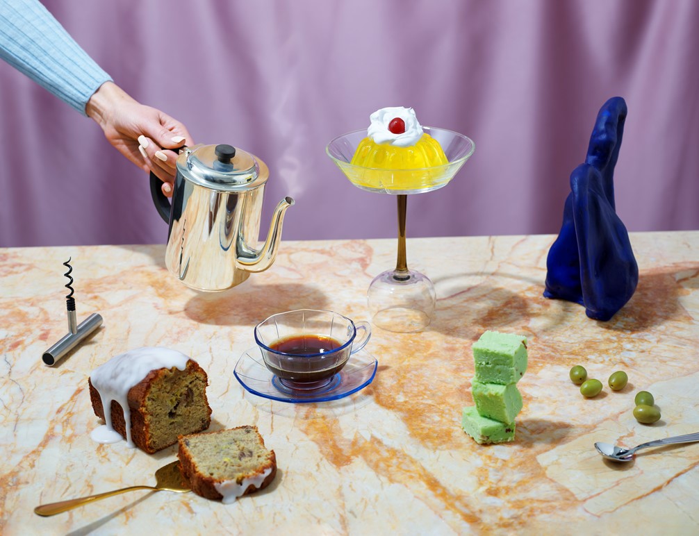 Hånd i lyseblå genser skjenker kaffe fra sølvkanne med formkake, oliven, vinåpner og gele dandert på marmorbord