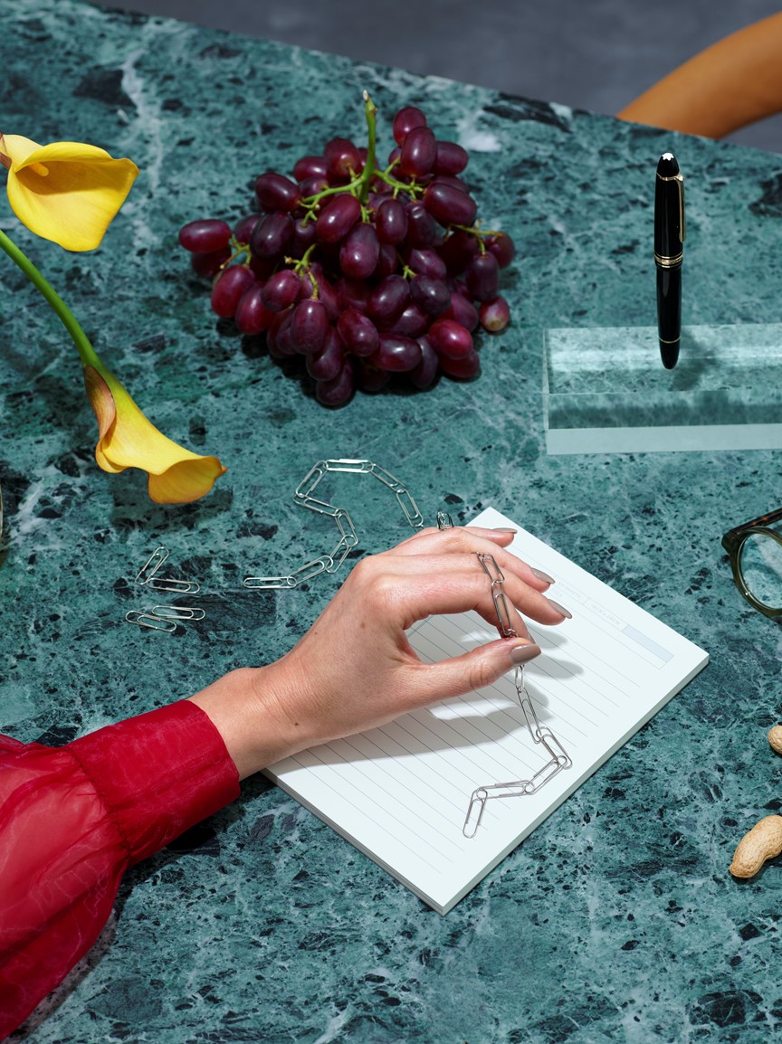 Hånd i rød bluse som holder lenke med binders over grønt marmorbord ved siden av gule blomster og druer
