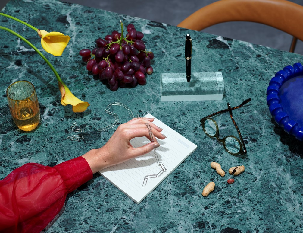 Hånd i rød bluse som holder lenke med binders over grønt marmorbord ved siden av gule blomster og druer